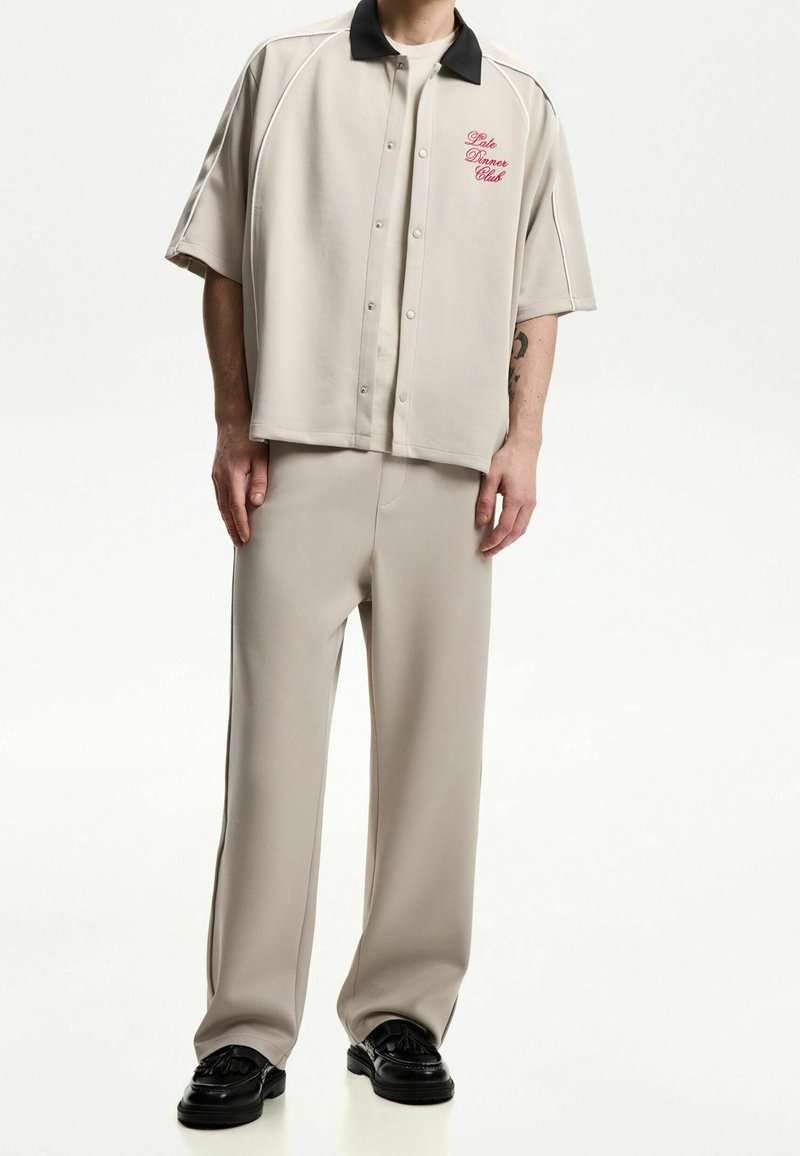 Béžová košile s krátkým rukávem na knoflíky s černým límcem a červenou výšivkou "Late Dinner Club", doplněná o béžové kalhoty a černé boty.