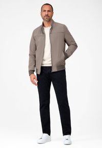 Brun bomberjacka med dragkedja, sidofickor och ribbade ärmar, bärs över en beigetröja och svarta jeans. Vita sneakers fulländar looken.