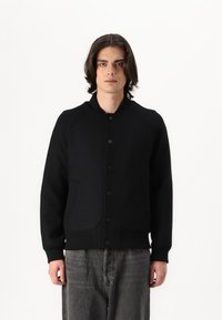 Svart bomberjacka tillverkad av ett mjukt, texturerat material. Har en ribbad krage, knappstängning och två framfickor, vilket skapar en strukturerad silhuett.