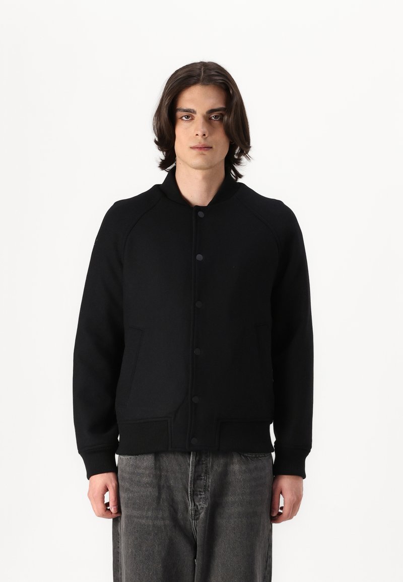 Svart bomberjacka tillverkad av ett mjukt, texturerat material. Har en ribbad krage, knappstängning och två framfickor, vilket skapar en strukturerad silhuett.