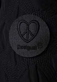 Černá prošívaná látka s kulatou vyšitou nášivkou srdce a symbolu míru, označená "Design". Texturovaný povrch s kontrastním stehem.