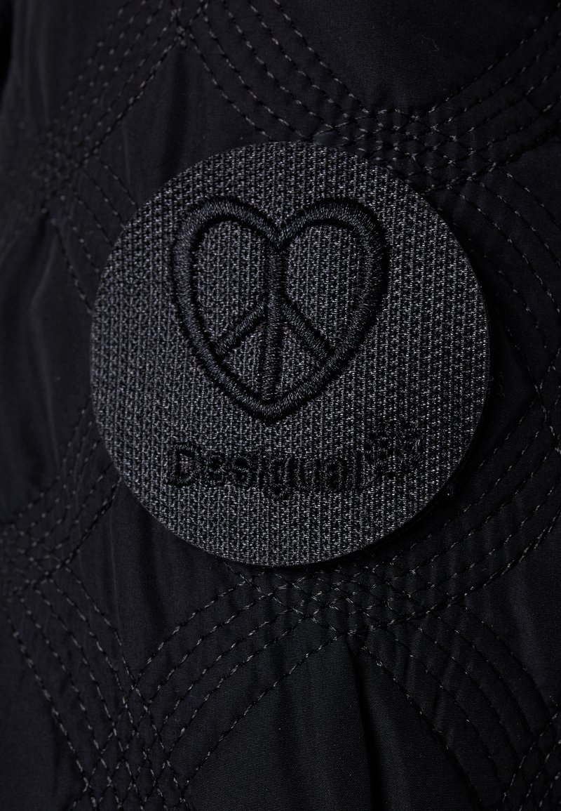 Černá prošívaná látka s kulatou vyšitou nášivkou srdce a symbolu míru, označená "Design". Texturovaný povrch s kontrastním stehem.