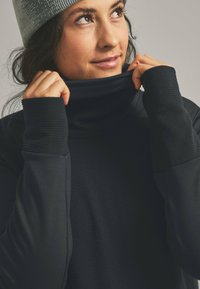 Schwarzes Langarmoberteil mit hohem Kragen, das über ein strukturiertes Strickdesign an den Bündchen und Schultern verfügt. Das Model hält den Kragen hoch und trägt eine hellgraue Mütze.