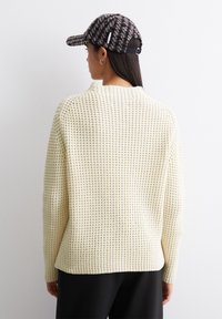 Cremfarbener Strickpullover mit strukturiertem Muster, lockerer Schnitt und überschnittenen Schultern. Kombiniert mit einer dunkel gemusterten Mütze.