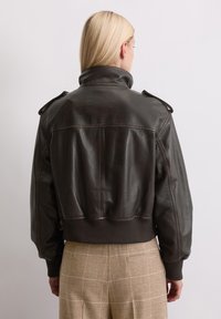 Braune Lederbomberjacke mit hohem Kragen, gerippten Bündchen und Schulterklappen, kombiniert mit karierter beigefarbener Hose.