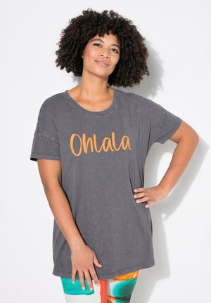 Šedé oversized tričko s kulatým výstřihem a krátkými rukávy, na kterém je nápis "Ohlala" v oranžové kurzívě. Kombinováno s barevnými legínami.