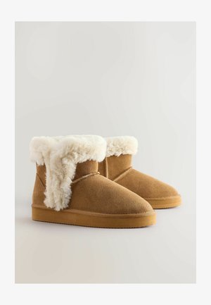 Botas de ante marrón hasta el tobillo con un puño de pelo sintético blanco y esponjoso, puntera redonda y suela de goma plana. Detalles de costura a lo largo de los lados.