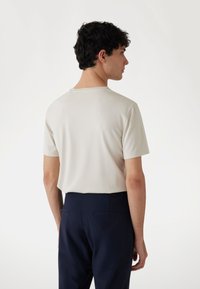 Helle beige Kurzarm-T-Shirt mit Rundhalsausschnitt. Glatte Textur, minimalistisches Design. Getragen mit dunkelblauen Hosen, Rückansicht wird gezeigt.