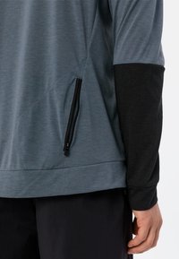 Top sportivo con corpo grigio e maniche lunghe nere, dotato di una tasca con zip sul lato. Realizzato in tessuto leggero e liscio.