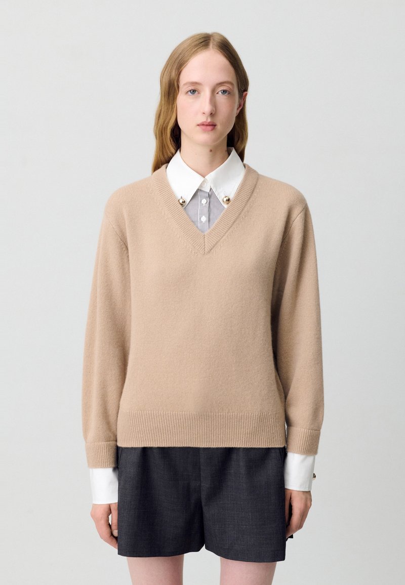 Pull beige en V, fabriqué en tissu tricot doux, avec des poignets et un ourlet côtelés. Porté sur une chemise blanche avec un col rayé et des embellissements ronds.