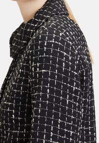 Czarna i biała, fakturowana kurtka z tweedu z szerokim kołnierzem, noszona przez osobę pokazaną od szyi do połowy torsu w profilu.