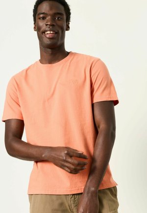 Lächender Mann in einem lachsfarbenen kurzärmligen T-Shirt und beigen Hosen, der seinen linken Ärmel vor einem schlichten hellen Hintergrund hochkrempelt.