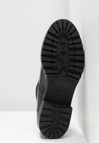 Semelle d'une botte noire avec des motifs de crampons profonds et "ANNA FIELD" embossé près du talon, photographiée sur un fond d'angle blanc.