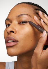 Brauner Eyeliner-Stift wird in der Nähe des Auges aufgetragen, mit natürlichem Hautton und minimalem Makeup. Heller Hintergrund, Fokus auf den Gesichtsmerkmalen.
