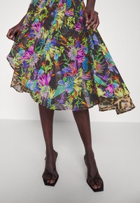Femme portant une jupe midi noire à fleurs tropicales colorées, associée à des sandales à talons noires à lanières.