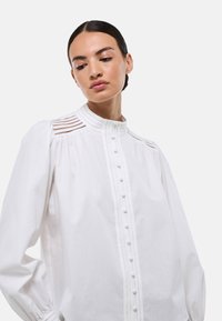 Blouse blanche en coton avec un col haut, un devant à boutons et des manches froncées. Présente des coutures décoratives et un subtil détail de texture.