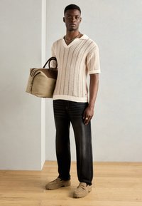 Gestricktes cremefarbenes Poloshirt mit V-Ausschnitt, schwarze Jeans und beige Slippers. Hält eine braune Tragetasche mit zweifarbigen Akzenten.