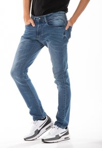 Jean en denim bleu ajusté avec un lavage moyen, design à cinq poches et détails de décoloration subtils, associé à des baskets blanches et grises.