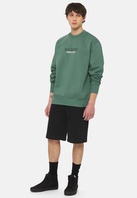 Grøn sweatshirt med ribbede manchetter og kant, der har teksten "DICKIES SINCE 1922". Kombineret med sorte shorts og sorte højhalsede sneakers.