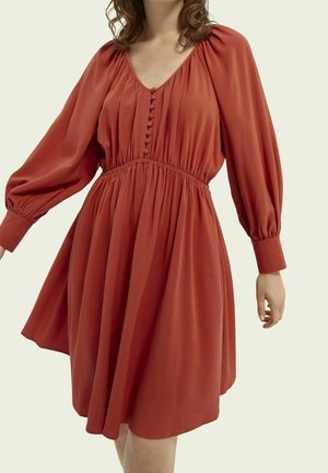 Okerkleurige jurk met lange pofmouwen, een getailleerde taille en knoopdetails op het lijfje, gedragen door een persoon met golvend haar.