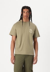 Olive green cotton t-shirt with short sleeves, round neckline, and white circular text "FREEDOM CHAOS" on the chest. Plain fabric texture.