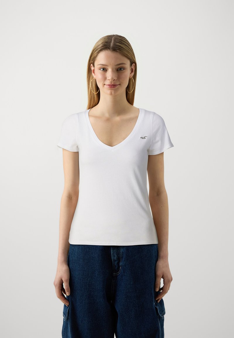 Biała bawełniana koszulka z głębokim dekoltem w kształcie V i krótkimi rękawami, ozdobiona małym szarym logo na piersi. Noszona z ciemnoniebieskimi, luźnymi dżinsami.