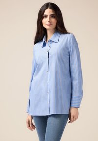 Camisa azul de botones con rayas verticales blancas y naranjas, de mangas largas y con cuello, hecha de una tela suave. Se lleva con jeans azules.