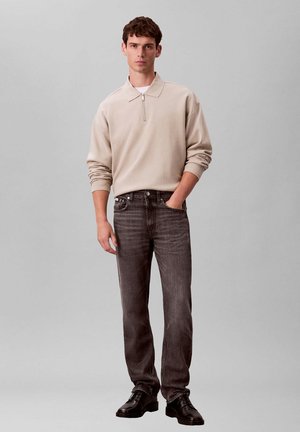 Giovane uomo in piedi su sfondo grigio indossa un maglione beige con collo con zip, jeans grigio scuro e scarpe in pelle nere.