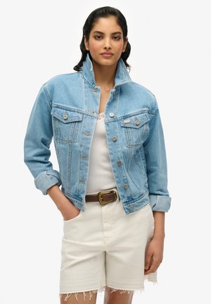 Chaqueta de mezclilla azul claro con bolsillos con solapa, herrajes plateados y un cuello alzado. Llevada sobre una camiseta blanca sin mangas y combinada con pantalones cortos blancos.