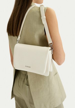 Kobieta w beżowej kamizelce bez rękawów i spodniach, niosąca białą, teksturowaną torebkę na ramię marki Lasocki z regulowanym paskiem na plecach.