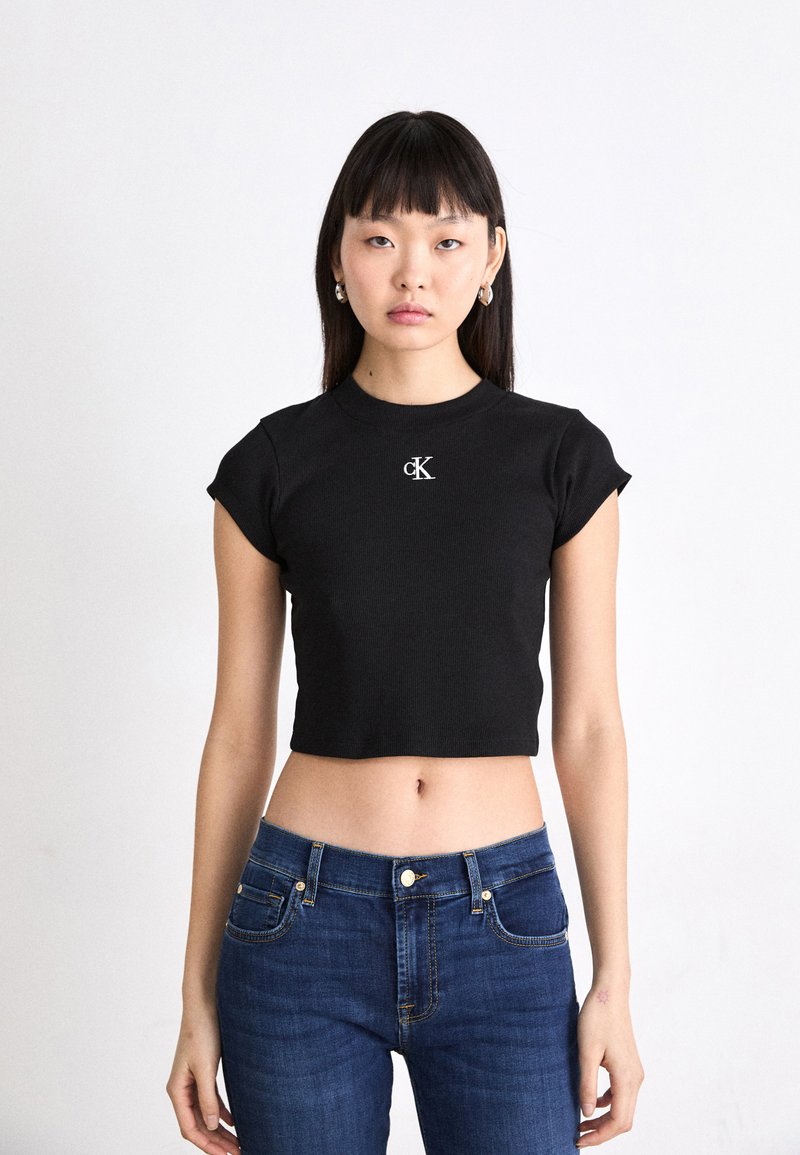 T-shirt noir court en tissu côtelé avec des manches courtes et un logo "CK" brodé sur la poitrine, associé à un jean fitted bleu profond.