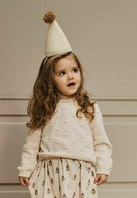 Lys beige strikkegenser med forhøyde bobler, kombinert med et mønstret kremfarget skjørt med små motiver. Barnet har på seg en kjegleformet lue med dusk.