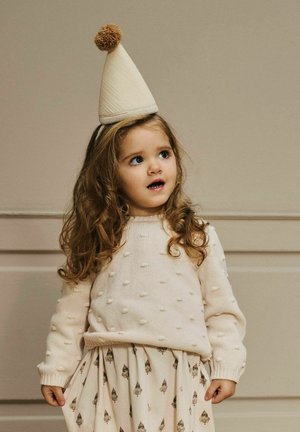 Svijetlo bež pleteni džemper s uzdignutim kuglicama, kombiniran s kremastom suknjom s uzorkom malih dizajna. Dijete nosi konusasti šešir s potpom.