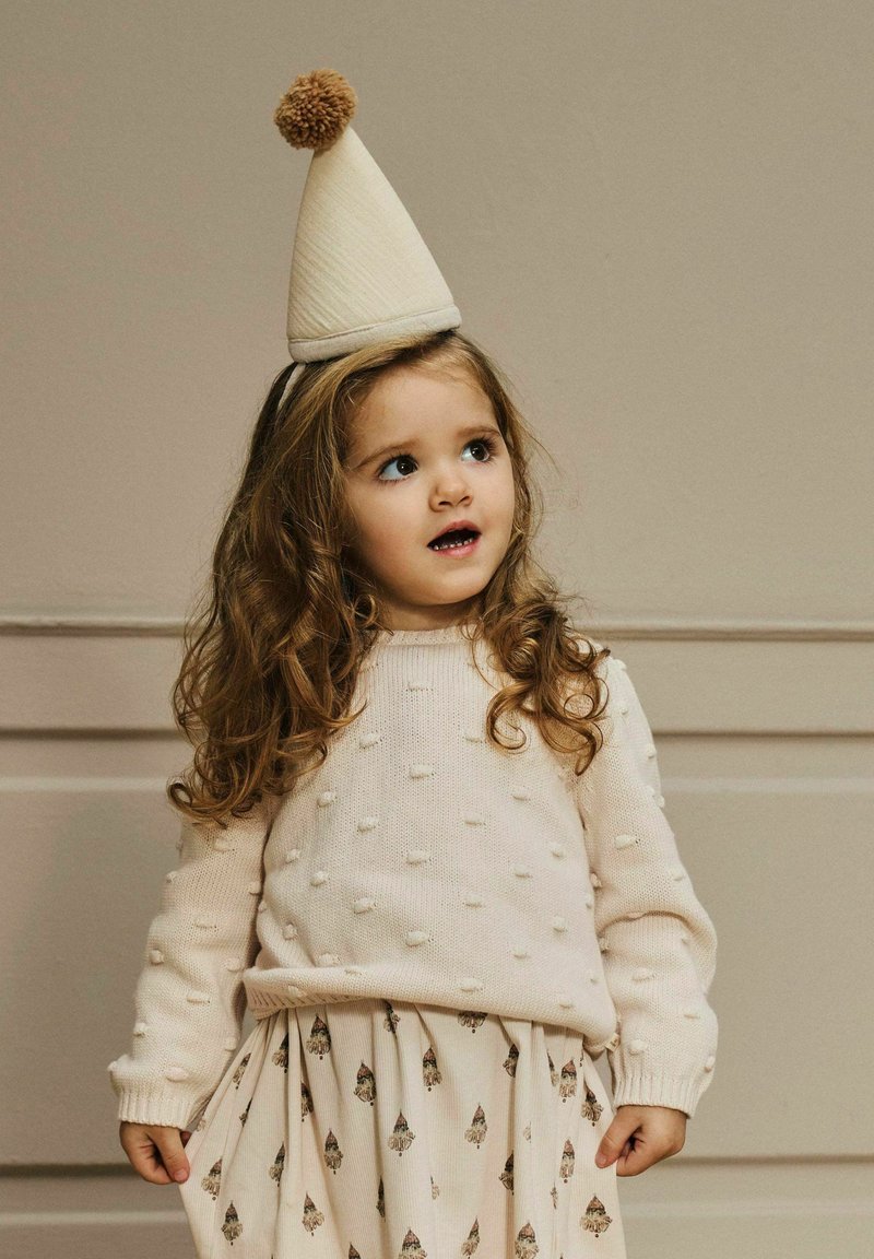 Lys beige strikkegenser med forhøyde bobler, kombinert med et mønstret kremfarget skjørt med små motiver. Barnet har på seg en kjegleformet lue med dusk.