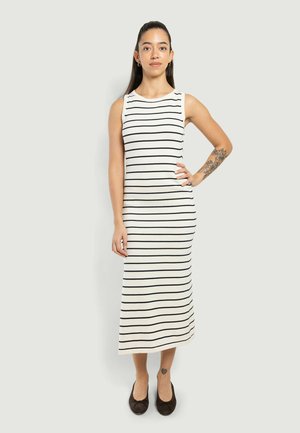 Donna con capelli lunghi e scuri in piedi che indossa un vestito bianco senza maniche con sottili strisce orizzontali nere, ballerine nere e tatuaggi floreali visibili sul braccio e sulla caviglia.