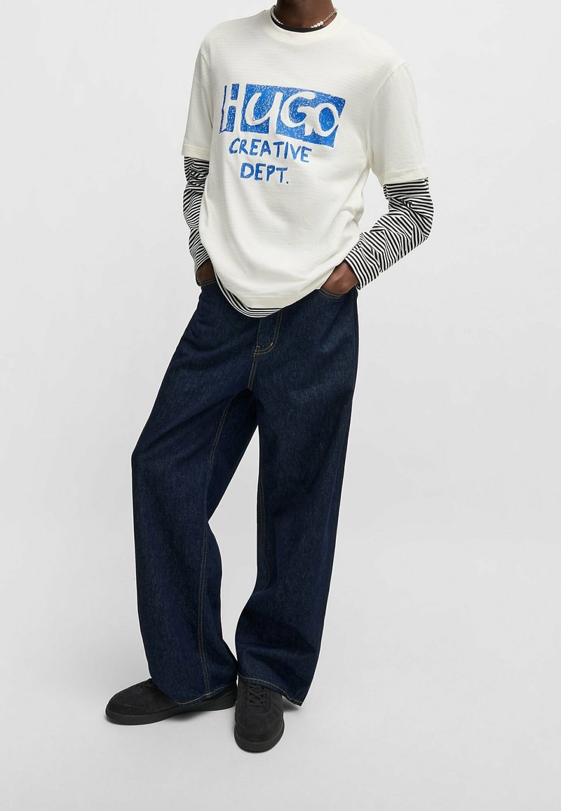Personne portant une chemise blanche à manches courtes avec le texte bleu « HUGO CREATIVE DEPT. » sur des manches longues rayées noir et blanc, un jean large foncé et des chaussures noires.