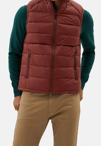 Bordeauxrote Steppweste mit horizontalen Nähten, seitlichen Reißverschlusstaschen und hohem Kragen, getragen über einem grünen Pullover. Strukturierter Stoff.