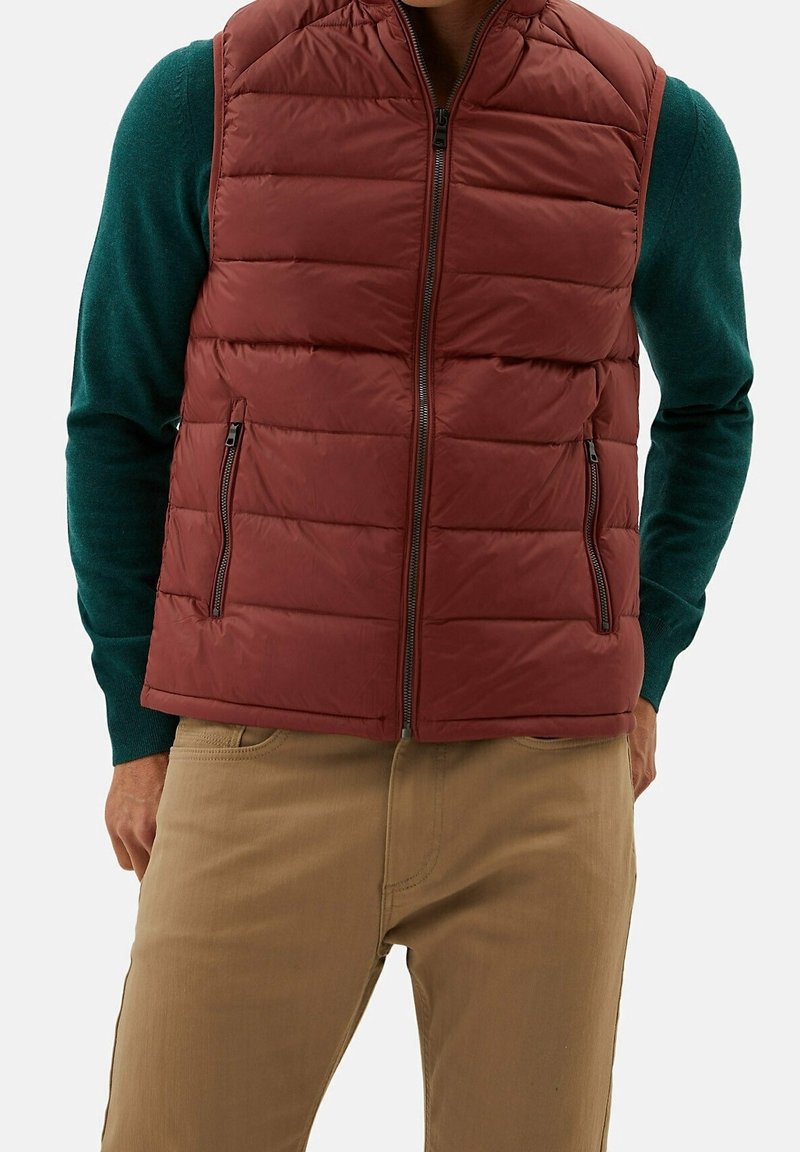 Bordeauxrote Steppweste mit horizontalen Nähten, seitlichen Reißverschlusstaschen und hohem Kragen, getragen über einem grünen Pullover. Strukturierter Stoff.