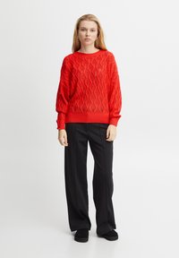 Pulover roșu tricotat cu un model în diamant, cu manșete și tiv ondulat. Asortat cu pantaloni negri cu picioare largi și pantofi negri. Design simplu.