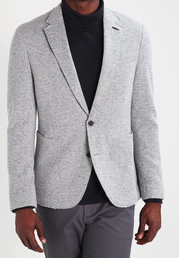 Hombre vistiendo un blazer gris claro sobre un suéter de cuello alto negro y pantalones gris oscuro, posando frente a un fondo blanco liso.