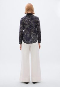 Blusa azul-marinho com padrão floral, mangas compridas e gola alta, combinada com calças brancas de pernas largas e sapatos pretos. Vista traseira.