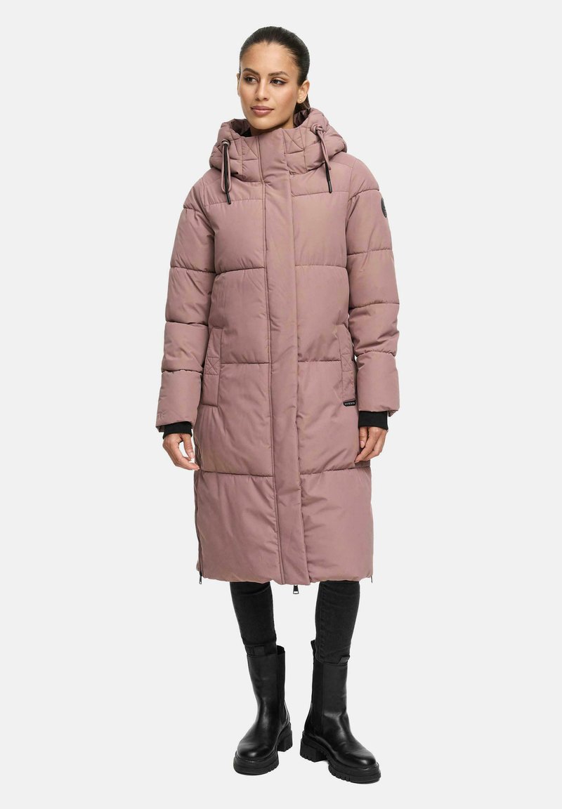 Kvinna som bär en lång mauvefärgad vadderad jacka med huva, svarta byxor och svarta klumpiga stövlar, stående mot en vit bakgrund.