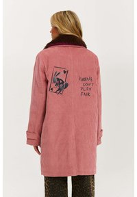 Ružový korduový kabát s hnedým umelým kožúškom. Na chrbte je grafika králika a text "srdcia TO NEROBIA SPRÁVNE." Dlhé rukávy a voľný strih.