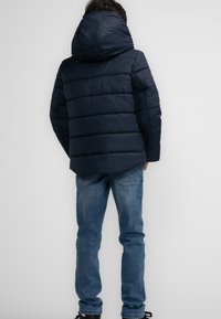 Marine pufferjack met een capuchon, met een gewatteerd ontwerp en horizontale stiksels. Draagt een paar blauwe jeans en is gezien van achteren.