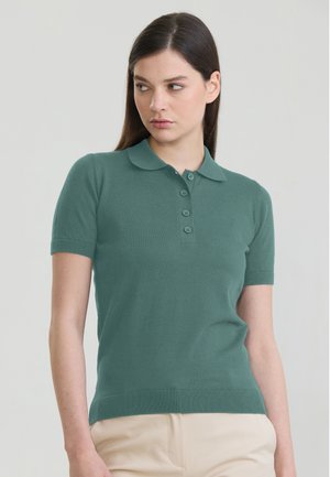 Kobieta z prostymi ciemnymi włosami, ubrana w zieloną koszulkę polo z krótkim rękawem i beżowe spodnie, lekko patrząca w prawo na jednolitym tle.