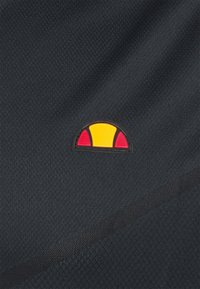 Le tissu texturé noir présente un petit logo en forme de demi-cercle, avec des sections jaunes et rouges. Le motif est cousu sur le matériau.