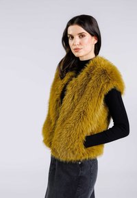Faux-Fur-Weste in Senfgelb mit dicken, strukturierten Fasern. Ärmelloses Design kombiniert mit einem enganliegenden schwarzen Rollkragenpullover und einem dunklen Rock.