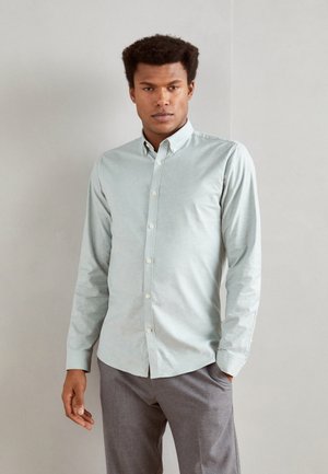 Camisa de manga larga verde claro con cuello abotonado, hecha de tela suave, con un corte ajustado y una textura sutil, combinada con pantalones grises.