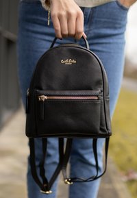 Černý kožený batoh s zaobleným tvarem, přední kapsou na zip, zlatými kovovými doplňky a nastavitelnými popruhy. Průhledné logo značky na vrchu.