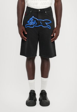 Shorts en denim noir avec un grand graphique bleu d'un chat, poches latérales et coupe décontractée ; associés à des chaussures noires et des chaussettes blanches.